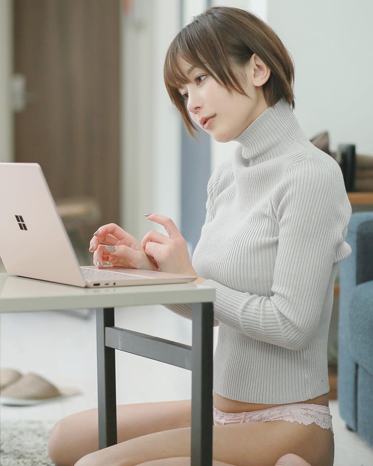 A young woman in a light gray turtleneck sweater sits on the floor, working on a laptop at a small table. She appears focused and engaged, with her hair styled in a short bob.
