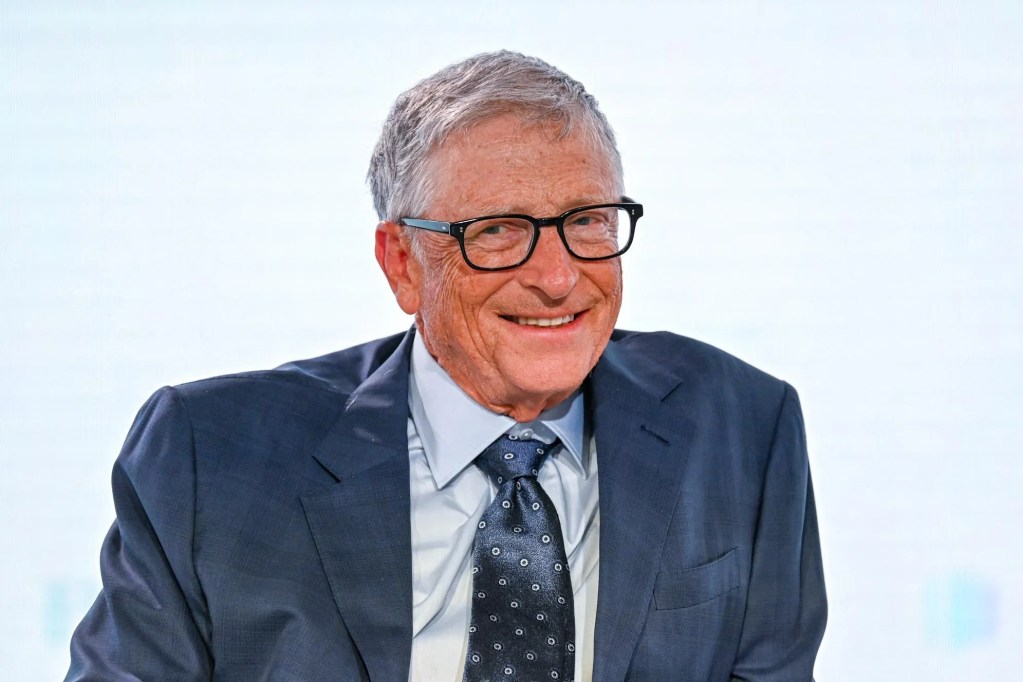 比爾·蓋茲微笑著穿著西裝,參加會議,背景為淺色牆面。