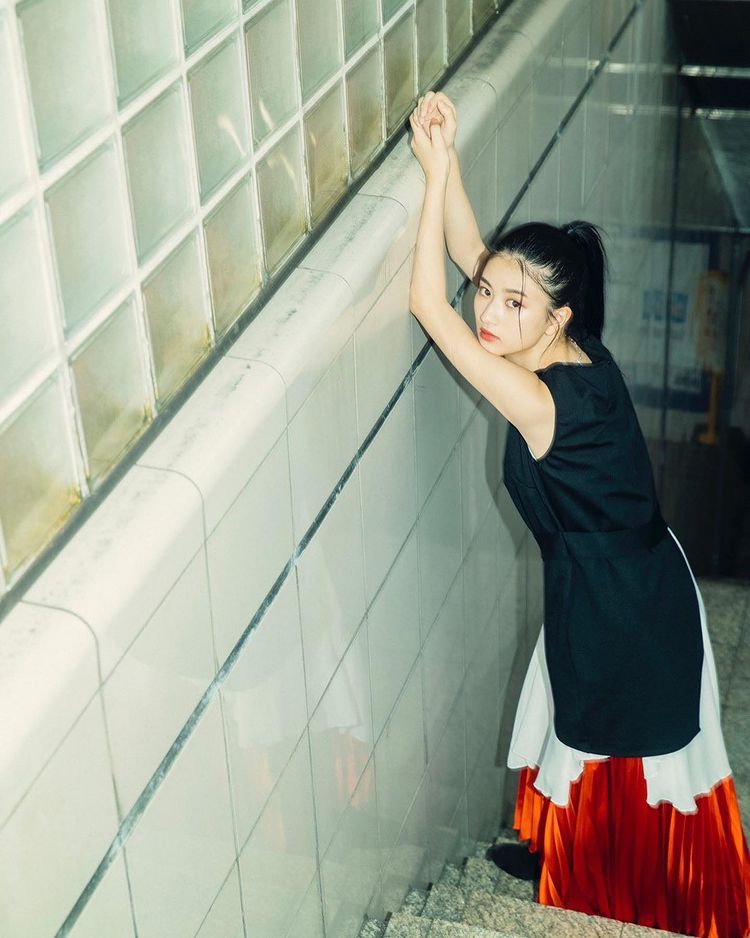 一位年輕女性站在樓梯旁，身穿黑色上衣和紅色裙子，雙手搭在牆邊，面帶淡淡的微笑，背景為潔白的瓷磚牆面
