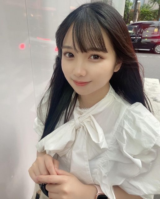 一位年輕女性站在城市街道上，穿著白色的襯衫，微笑著看向鏡頭，背景有模糊的車輛和街道景象。