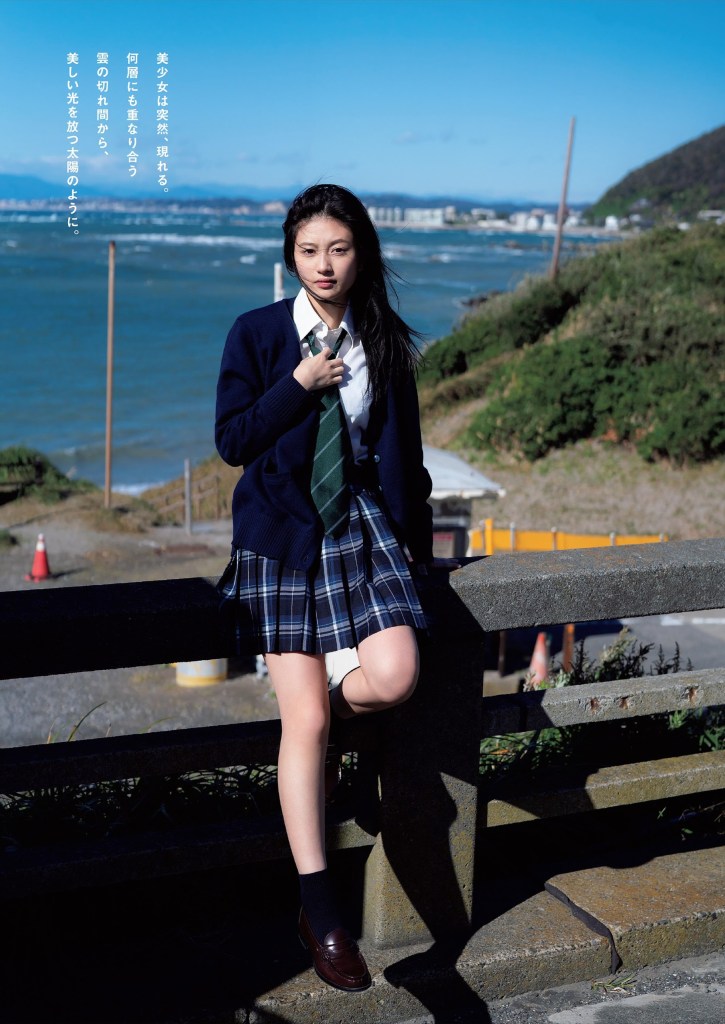 一位少女穿著學校制服，站在海邊的圍欄旁，背景是藍色的天空和波濤洶湧的海洋。