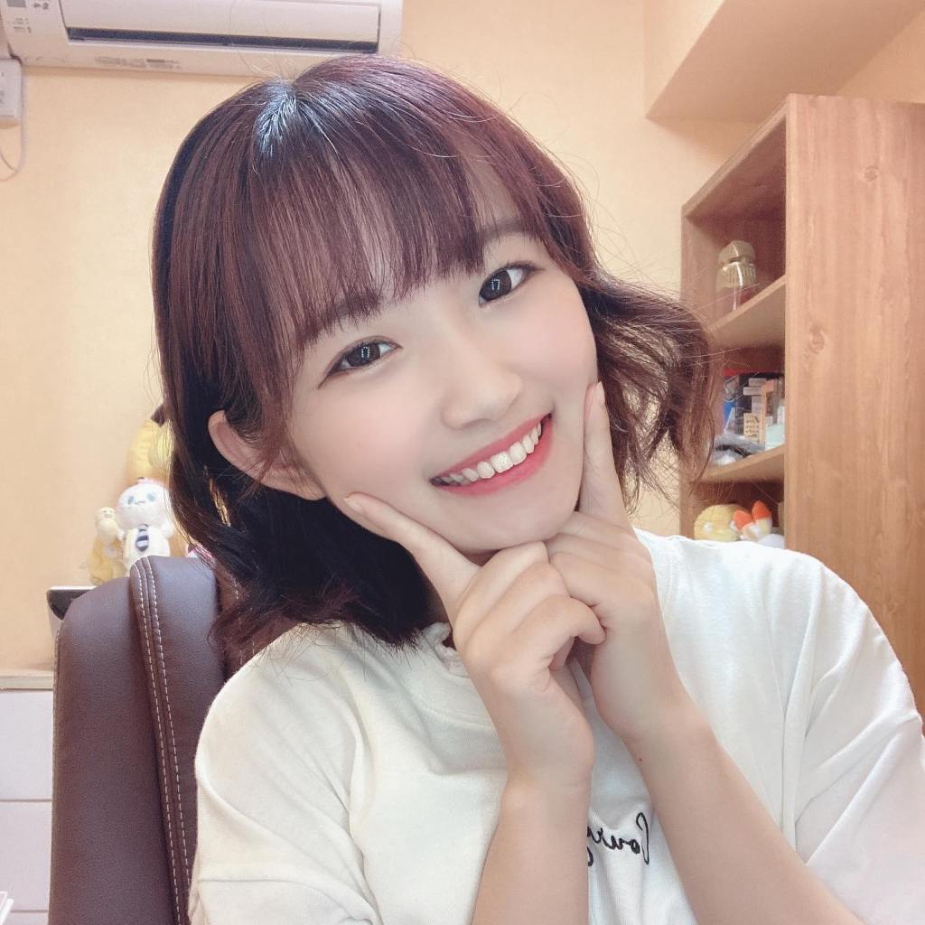 一位年輕女孩微笑著，雙手托著下巴，坐在辦公椅上，背景有桌子和書架，氣氛溫馨自然。