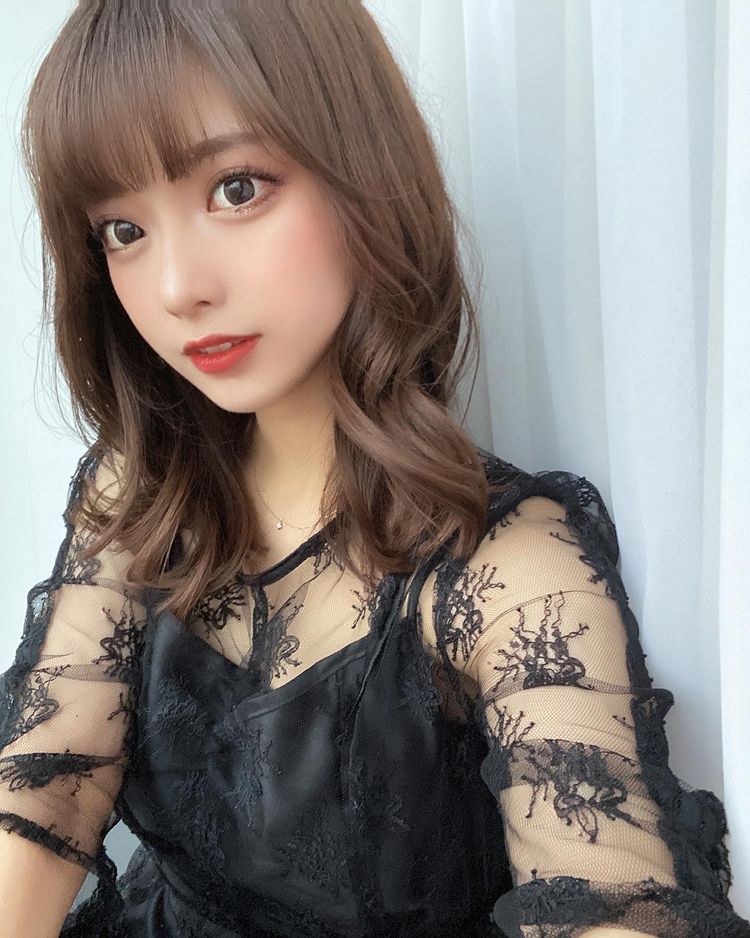 一位穿著黑色蕾絲上衣的年輕女性，面對鏡頭微笑，長髮及肩，背景為淺色窗簾。