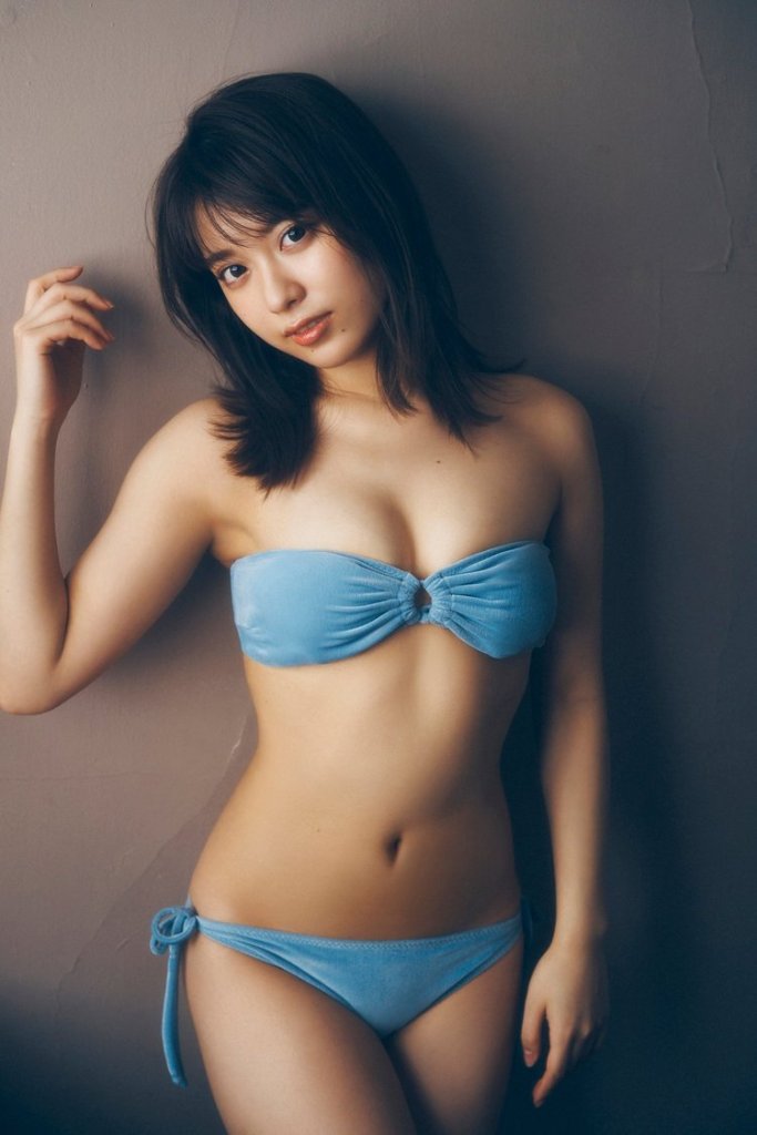 一位女性穿著藍色泳衣，站在牆邊微笑，手撐在頭髮旁，展現出自信的姿態。