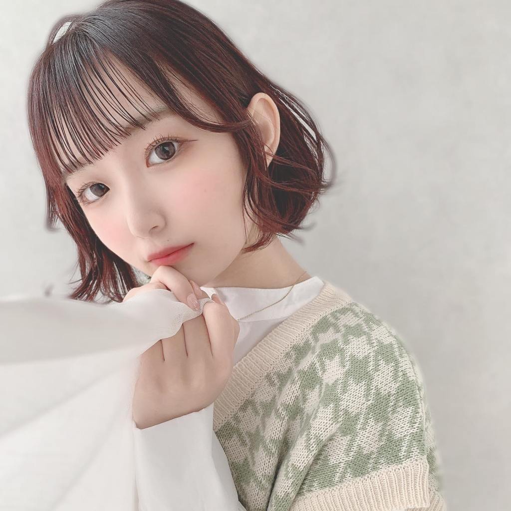 一位年輕女孩，擁有短髮和柔和的妝容，穿著綠色花紋的針織毛衣，輕輕握住白色面料，面帶微笑，背景模糊，營造出柔和的氛圍。