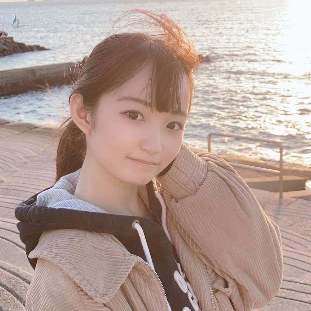 一位年輕女孩在海邊微笑，長髮隨風擺動，穿著棕色外套，背景為波光粼粼的海面。