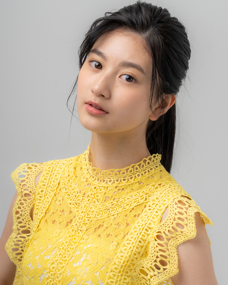 A young woman with long dark hair, wearing a vibrant yellow lace top, poses against a light grey background.