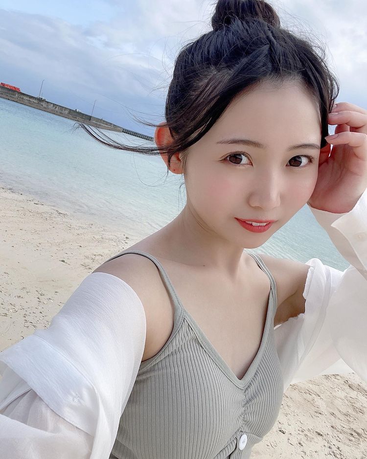 一位年輕女性在海邊拍攝的自拍，穿著淺綠色的無袖上衣和白色外套，背景為沙灘和海水，天氣晴朗。