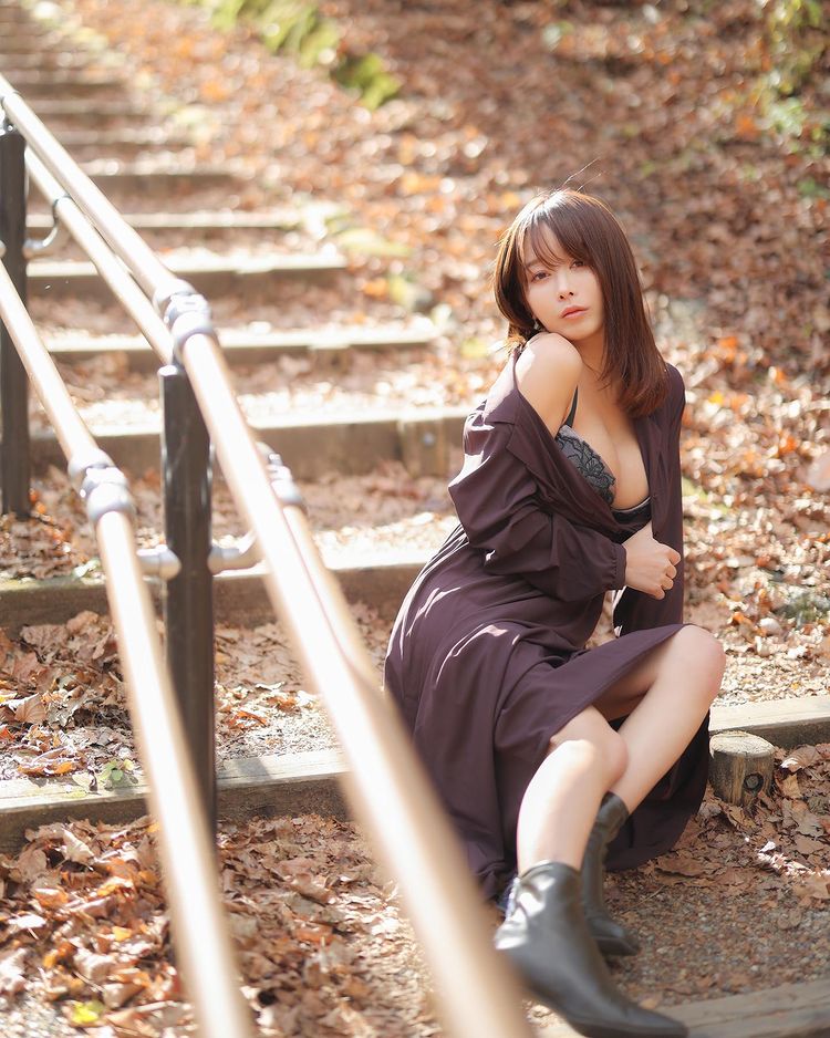 A woman sitting on stone steps surrounded by autumn leaves, wearing a black lace top and a long dark coat, with a contemplative expression.