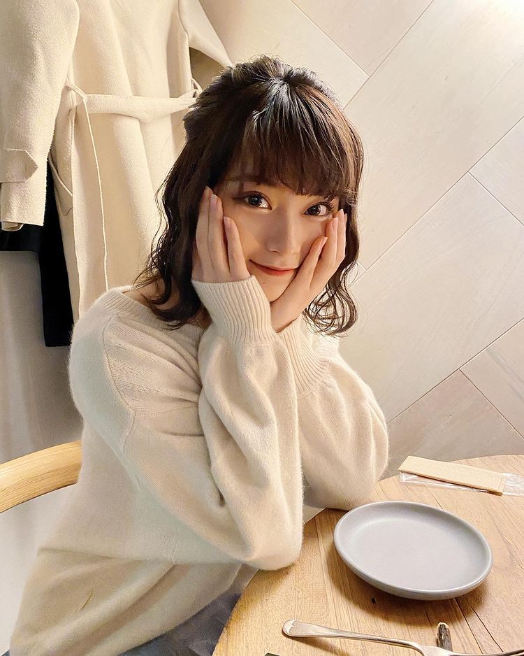 一位柔和微笑的女性，雙手托著臉頰，坐在木桌前，身穿淺色毛衣，背景是淺色牆壁和餐具。