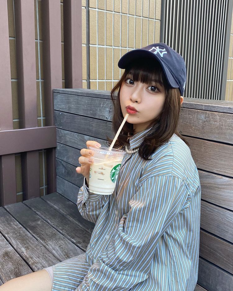 一位年輕女性坐在木椅上，穿著條紋襯衫，佩戴棒球帽，手中拿著一杯星巴克飲料，面帶微笑。