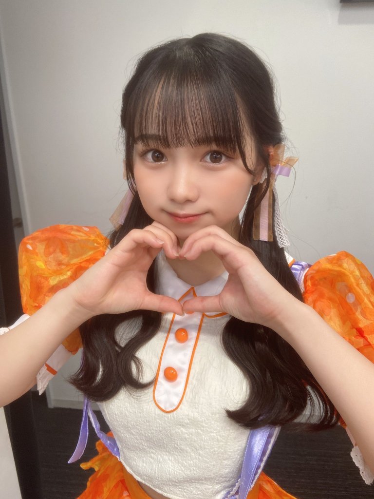 一名穿著橙色裝飾服的女孩，雙手做出愛心手勢，面帶微笑，展現出可愛的形象。