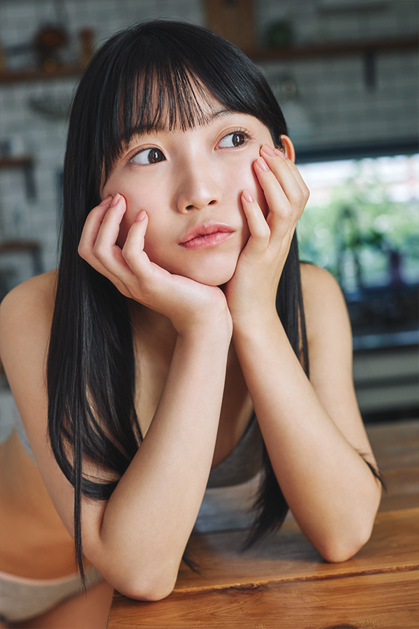 A young girl resting her chin on her hands, gazing thoughtfully with a slight pout. She has long black hair and is seated at a wooden table, with a soft-focus background showing greenery.