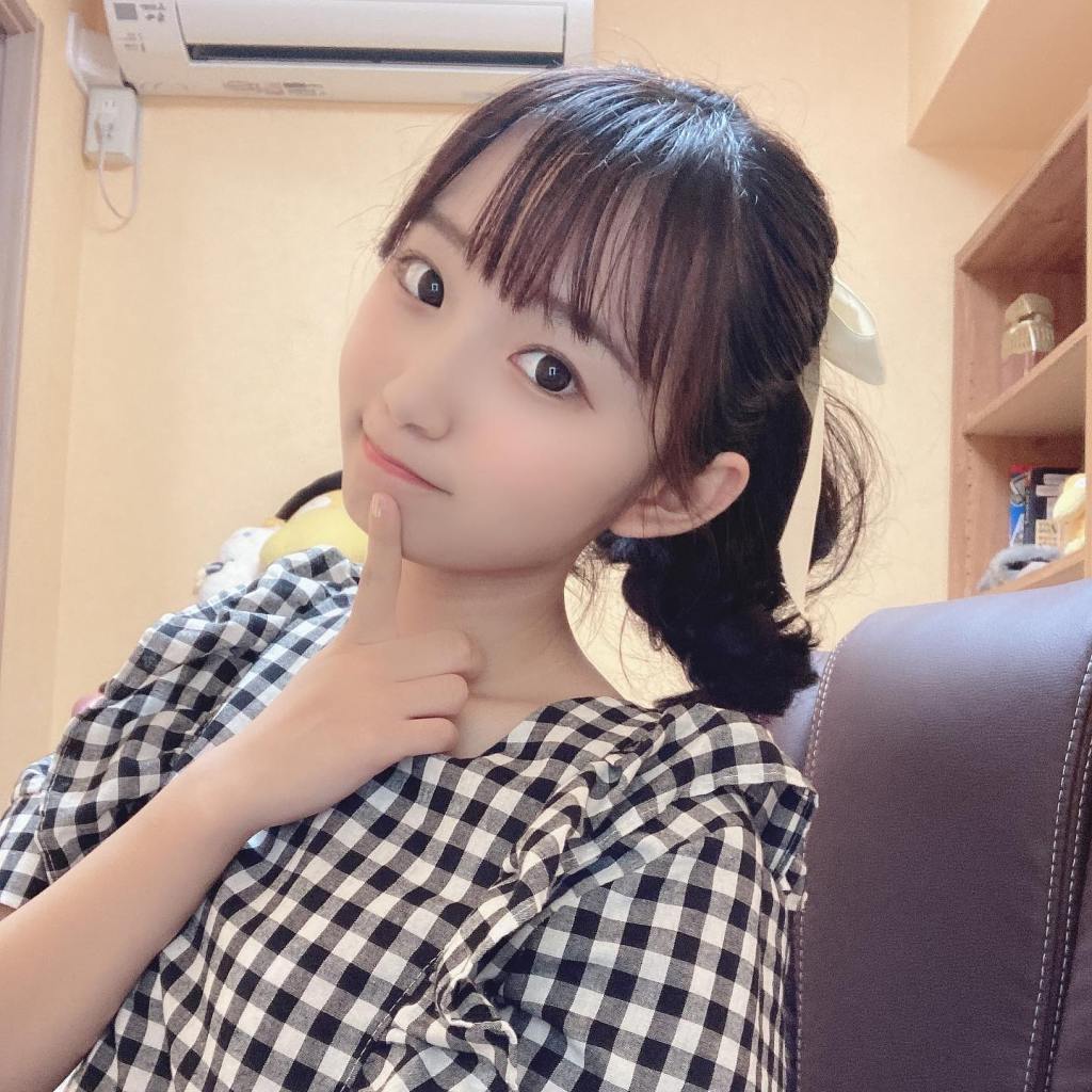 一位穿著格子衣服的女孩微笑著，手指放在臉頰旁，背景為室內環境。