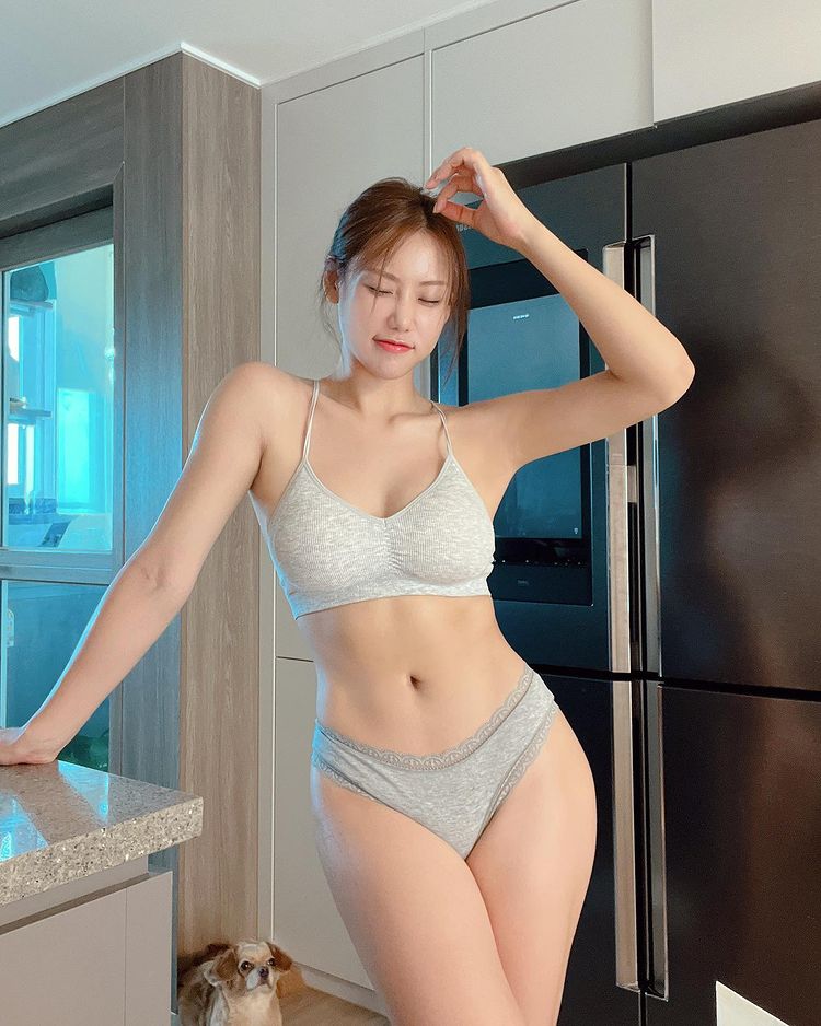 一位穿著灰色內衣的女性，站在廚房中微笑，手輕撫頭髮，身旁有一隻小狗。