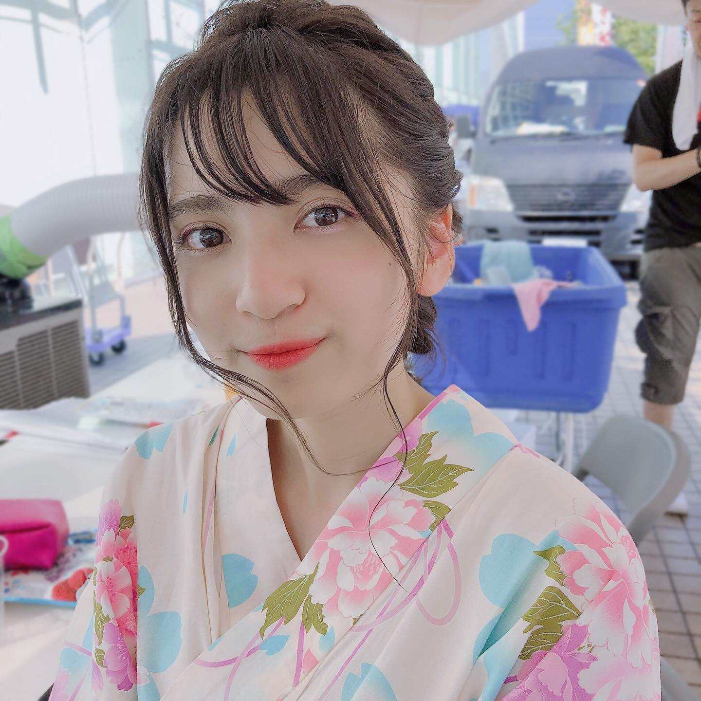 一位穿著花卉圖案和服的年輕女性，自信地微笑，背景有模糊的車輛和其他物品。