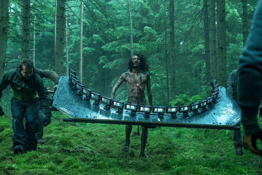 一群工作人員在樹林中搬運一個大型拍攝裝置，裝置上連接著多台iPhone，並顯示出一名滿身泥土的演員。