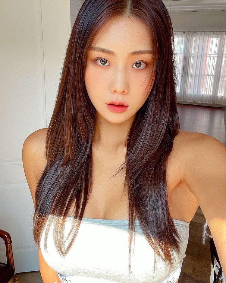 一位擁有長直髮的年輕女性，身穿緊身上衣，面對鏡頭微笑，背景為明亮的室內空間。