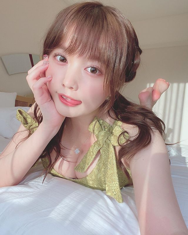 清純可愛的女性躺在床上，穿著綠色衣服，表情甜美，長髮及肩，手撫著臉頰，背景為明亮的室內環境