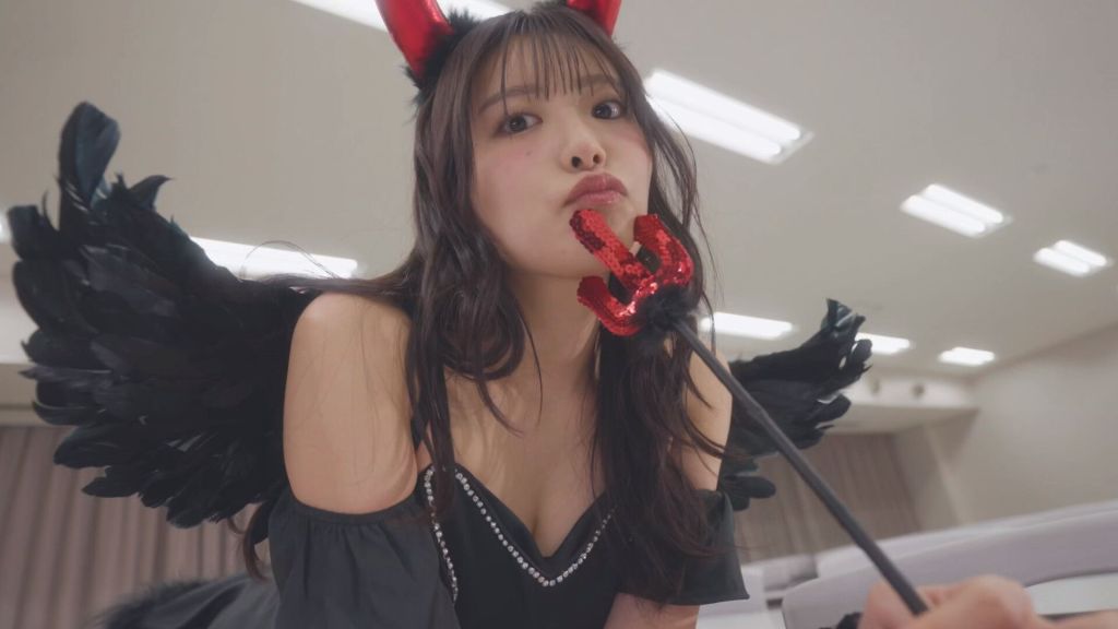 一名穿着黑色服装的女性，头戴红色小恶魔角，背后有黑色羽毛翅膀，嘴中叼着一根带有红色装饰的小恶魔叉，摆出俏皮的姿势。