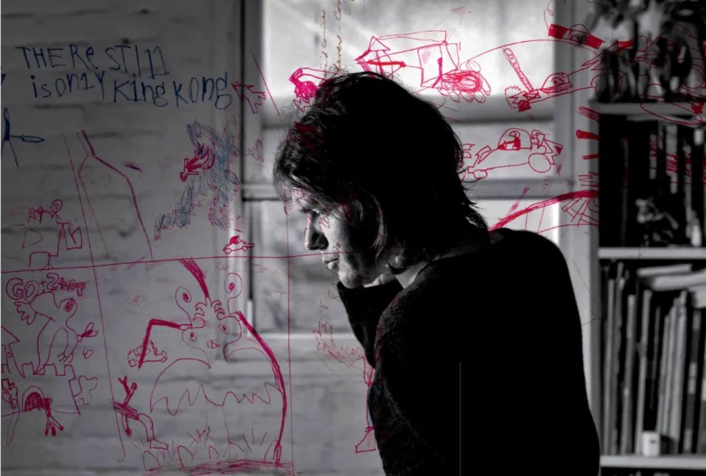 一位漫畫家側臉肖像，身後是插畫和文字的草圖，背景為模糊的室內環境。