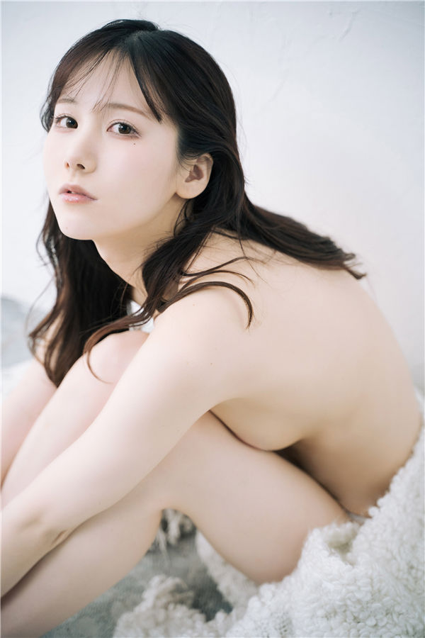 A close-up portrait of a young woman with long hair sitting against a white wall, looking contemplative while wrapped in a soft, fuzzy blanket.