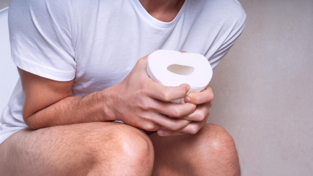 A person sitting on a toilet holding a roll of toilet paper, appearing to be experiencing discomfort.