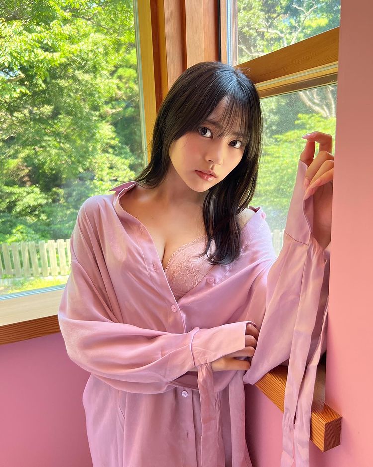 一位穿著淡紫色衬衫的女性，靠在窗边，微微侧身望向镜头，背景是綠色的自然景色。