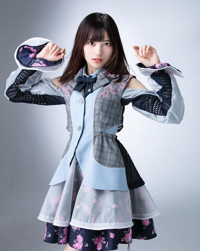 A young woman posing confidently in a stylish outfit featuring a checked jacket with floral accents and a layered skirt.