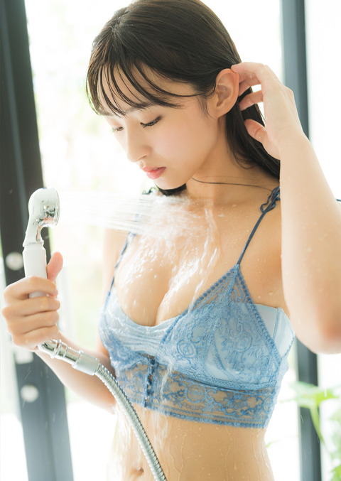A young woman with long hair is standing under a shower, gently running her fingers through her hair while water sprays down. She is wearing a light blue lace top and has a serene expression.