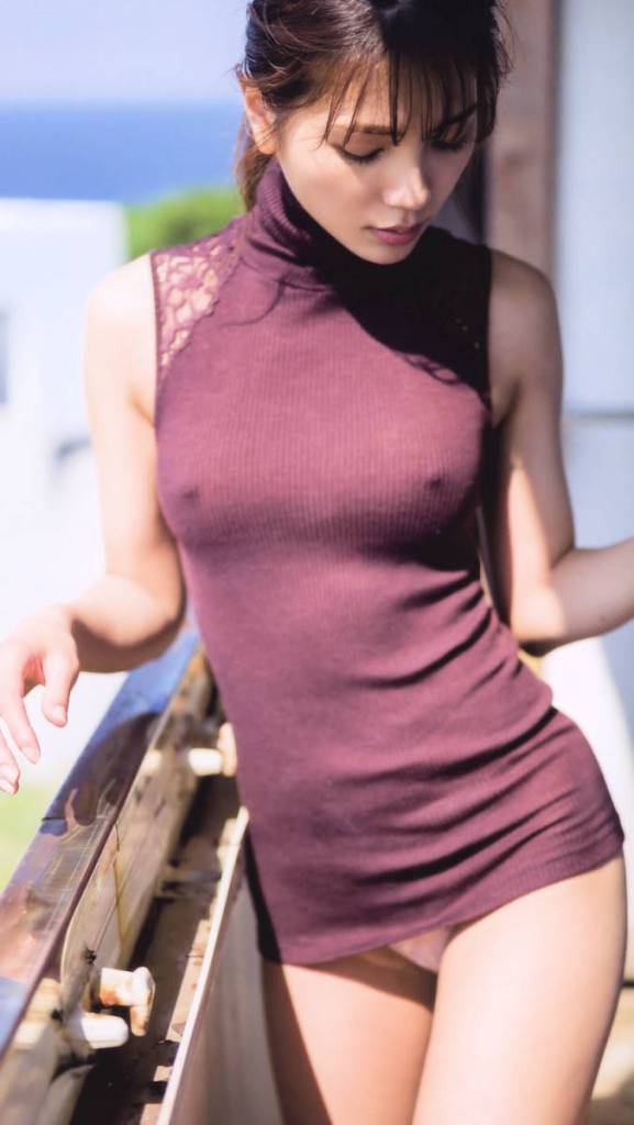A woman in a fitted, sleeveless turtleneck top stands by a railing, gazing downward with a serene expression. The background features a blurred view of greenery and the ocean.