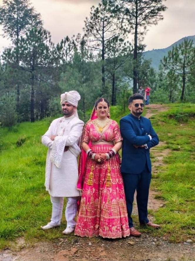 兩位身穿傳統服飾的男性與一位穿著靚麗紅色婚禮服的女性站在一起，背景為綠色的草地和樹林。