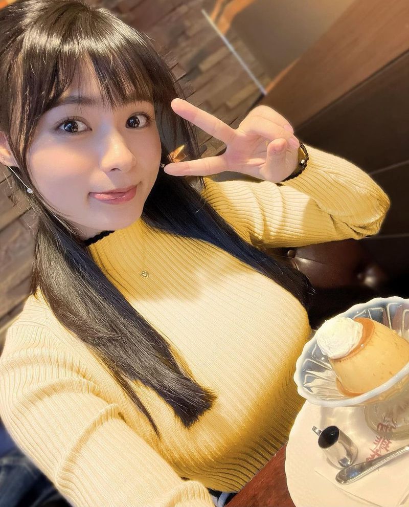 一位年輕女子穿著黃色毛衣，微笑並比著和平手勢，身旁有一碗甜品，背景為咖啡廳的牆面。