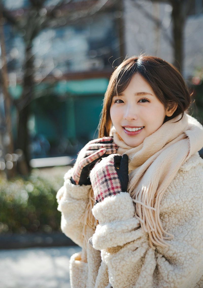 A young woman smiling warmly while wearing a fluffy coat, scarf, and gloves in a sunny outdoor setting.