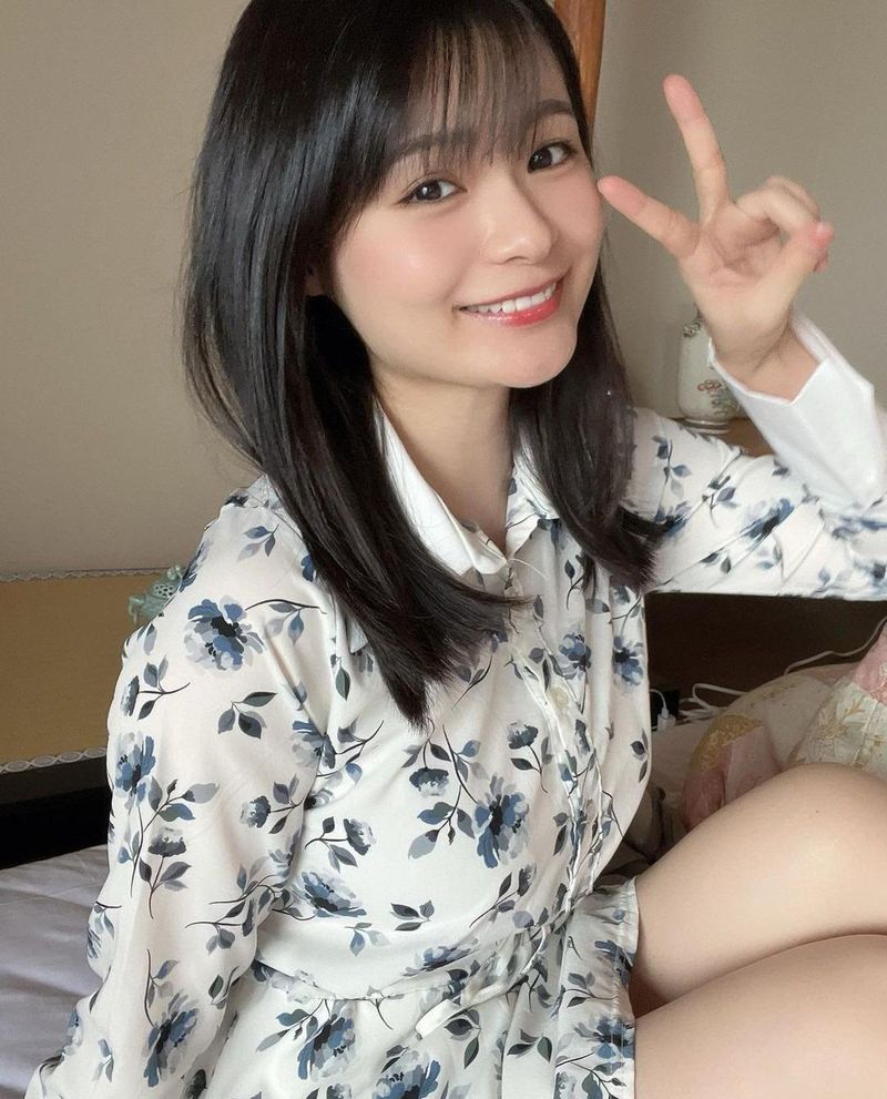 微笑著的女性，穿著花紋襯衫，手勢比出 V 字，坐在床上，背景柔和，給人一種可愛的氛圍。