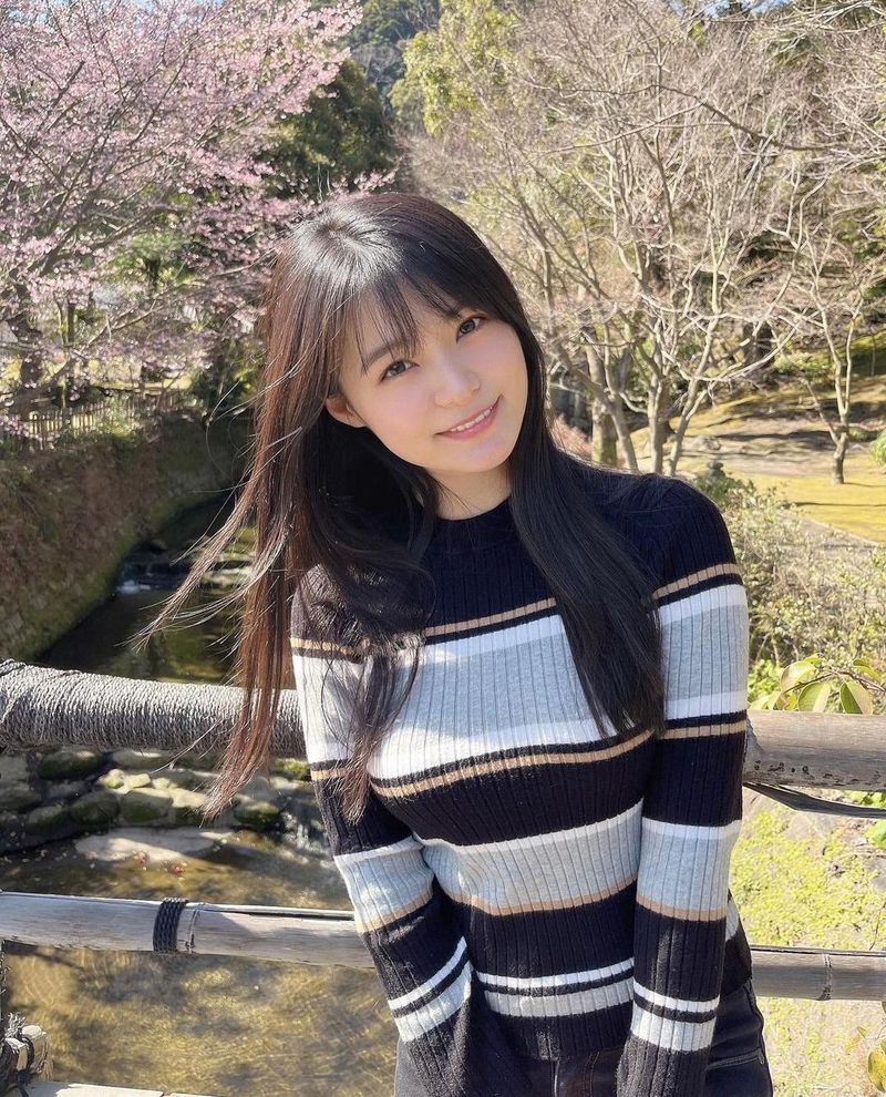 一名年轻女性在樱花树下微笑，穿着条纹毛衣，背景中有清澈的小溪和春天的自然景色。