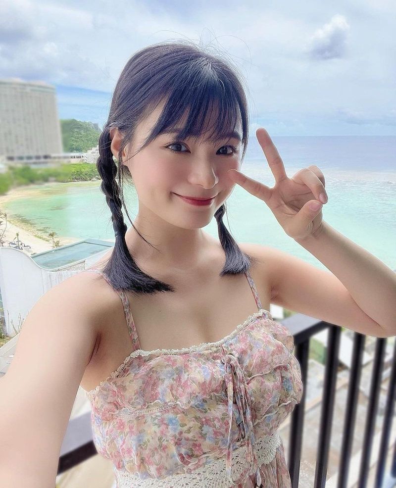 一名年輕女性在海邊的陽台上微笑，手勢比出和平的V字型，穿著花卉圖案的連衣裙，背景是海洋和藍天。