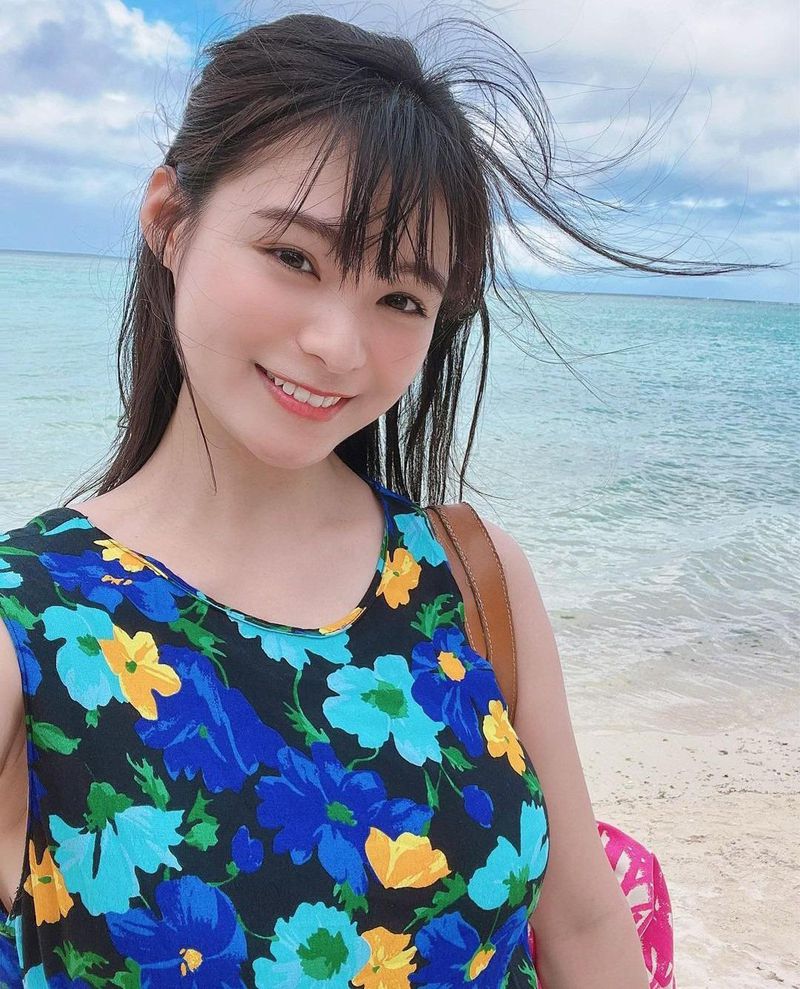 一位女性在海邊微笑著自拍，穿著花卉圖案的上衣，背景是藍色的大海和晴朗的天空。