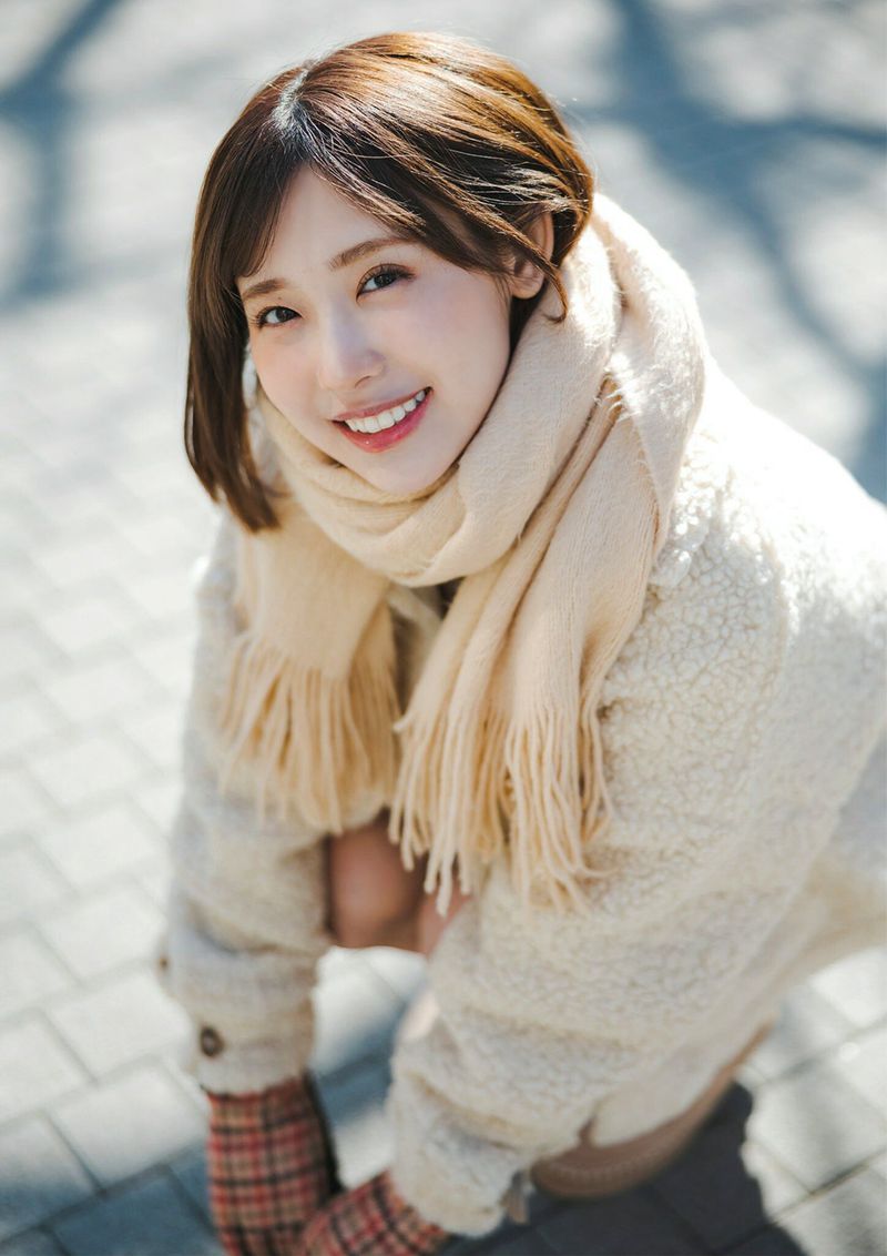 A young woman with a warm smile, wearing a beige scarf and a fluffy coat, crouched on a cobblestone path, with sunlight illuminating her face.