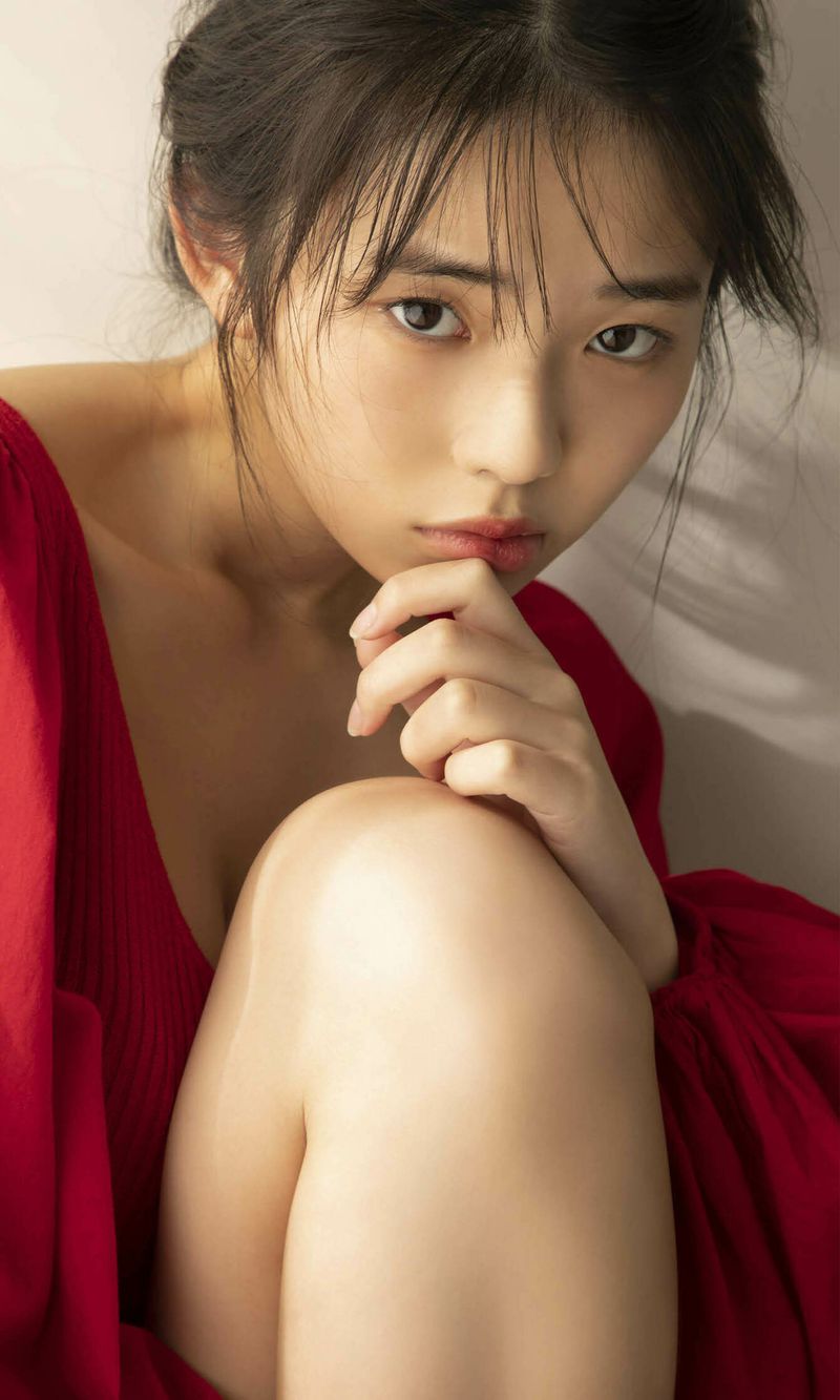 A young woman with dark hair, wearing a red top, gazes directly at the camera while resting her chin on her hand and hugging her knees.