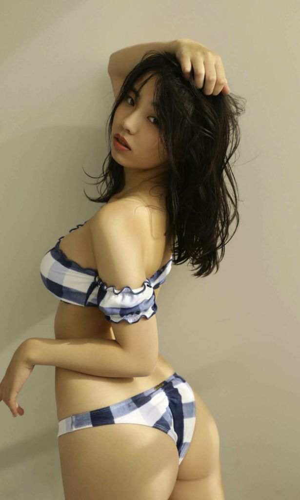 一位女性侧身站立，穿着蓝白格子泳衣，手抚摸着头发，背靠着一面光滑的墙面，表情自然。