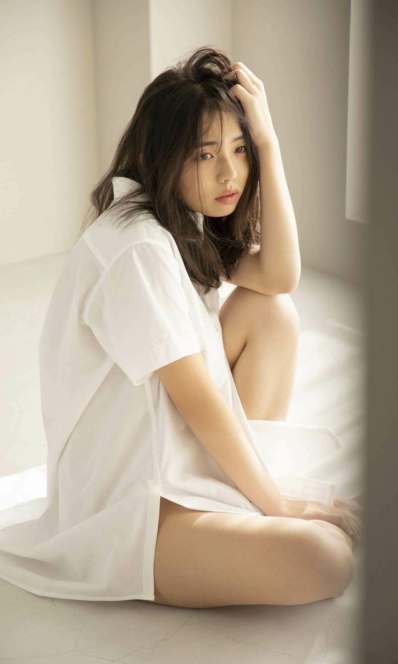 A young woman sitting on the floor, wearing a loose white shirt, with a contemplative expression and slightly tousled hair.