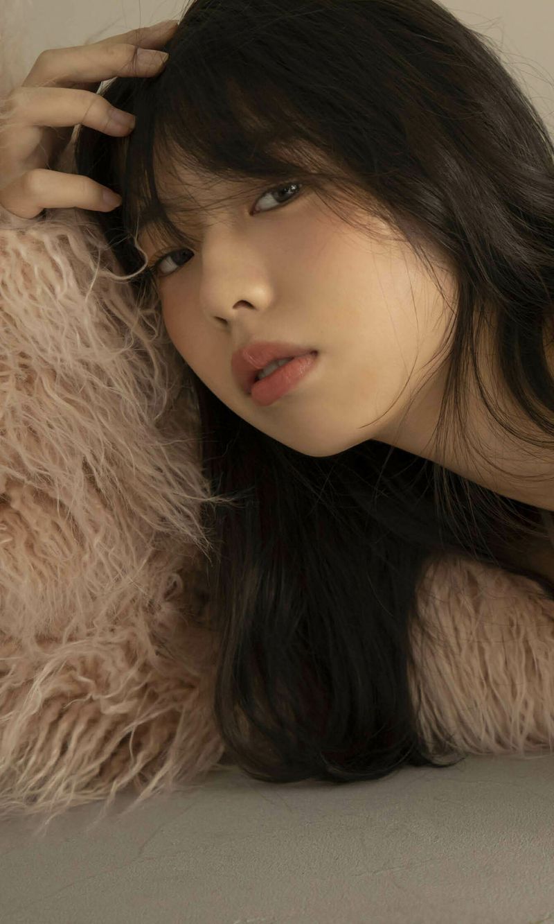 A close-up portrait of a young woman with long, dark hair, resting her head against a fluffy, light-colored pillow. She has a soft expression and appears thoughtful.