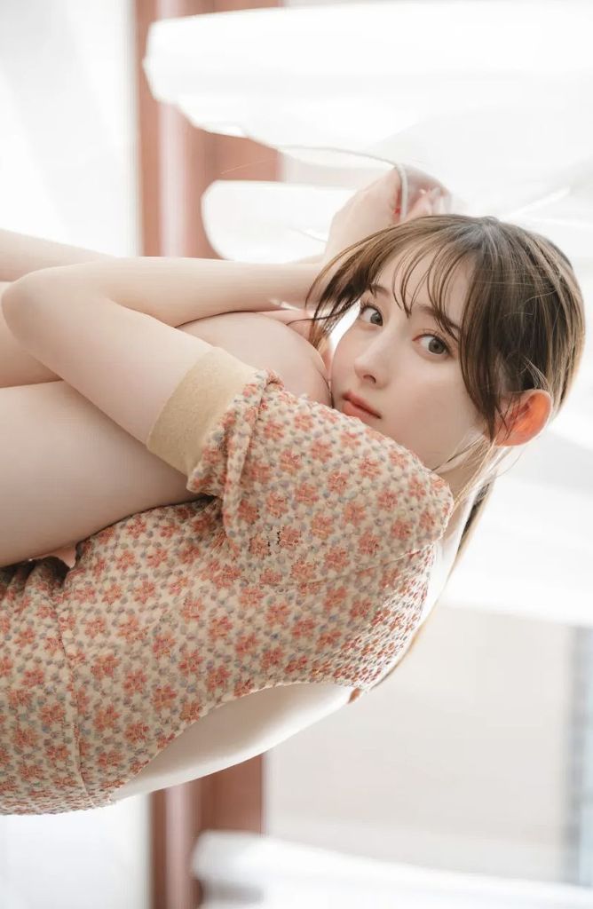A young woman with long hair is gracefully posed while sitting on a window ledge, wearing a floral patterned dress. She looks directly at the camera with a soft expression.