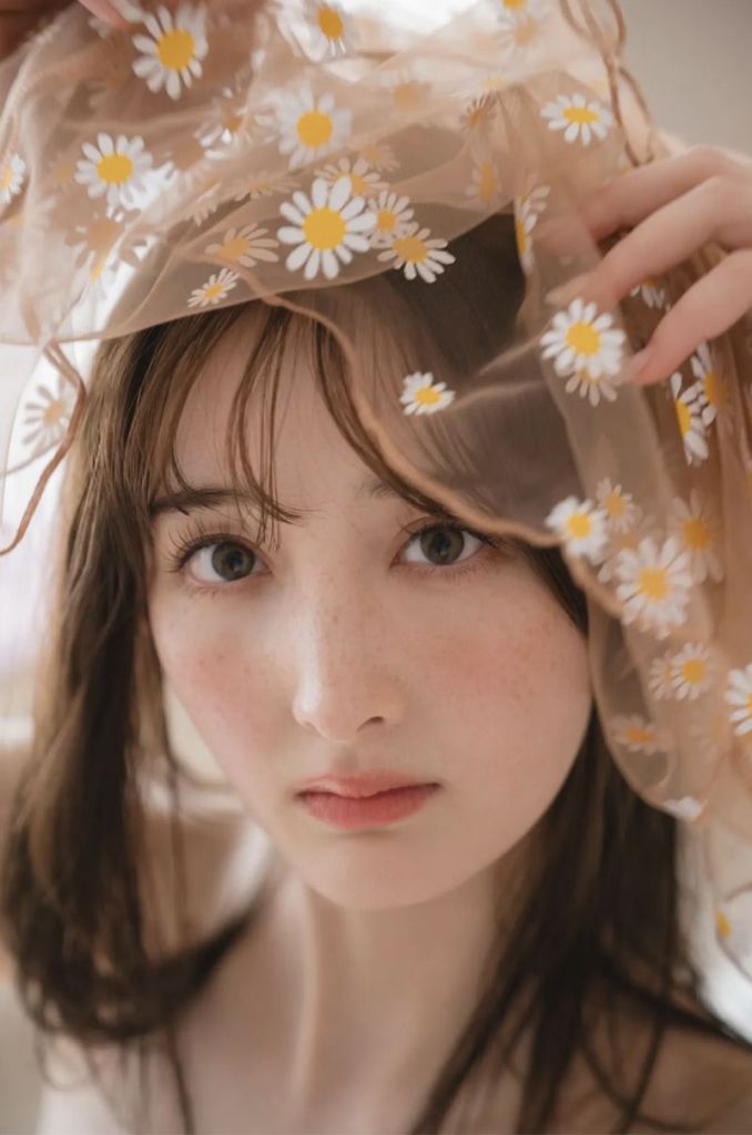 A close-up portrait of a young woman with long brown hair, wearing a sheer scarf adorned with daisies, gazing directly at the camera.