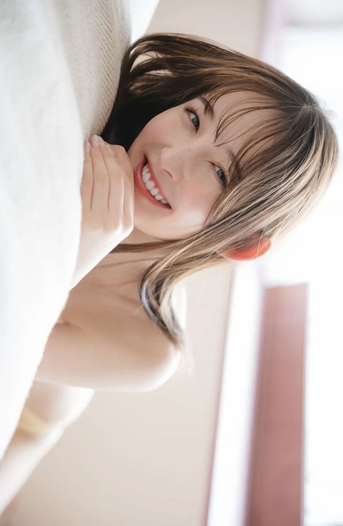 A smiling young woman with long hair, resting her chin on her hands while lying on a light-colored surface.