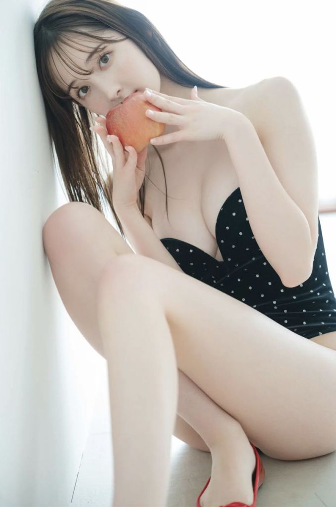 A woman in a polka dot swimsuit sitting against a wall, holding an apple close to her face.