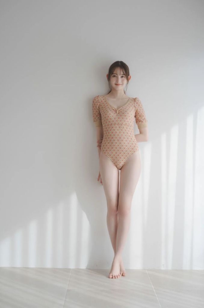A young woman posing in a patterned one-piece swimsuit against a light-colored wall, with soft natural lighting and shadows on the floor.