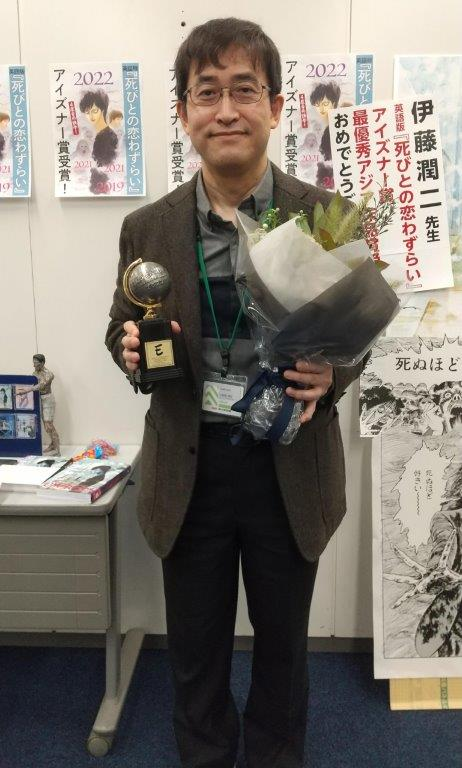 伊藤潤二獲得艾斯納獎的典禮上，手持獎盃和花束，笑容滿面，背景有漫畫海報