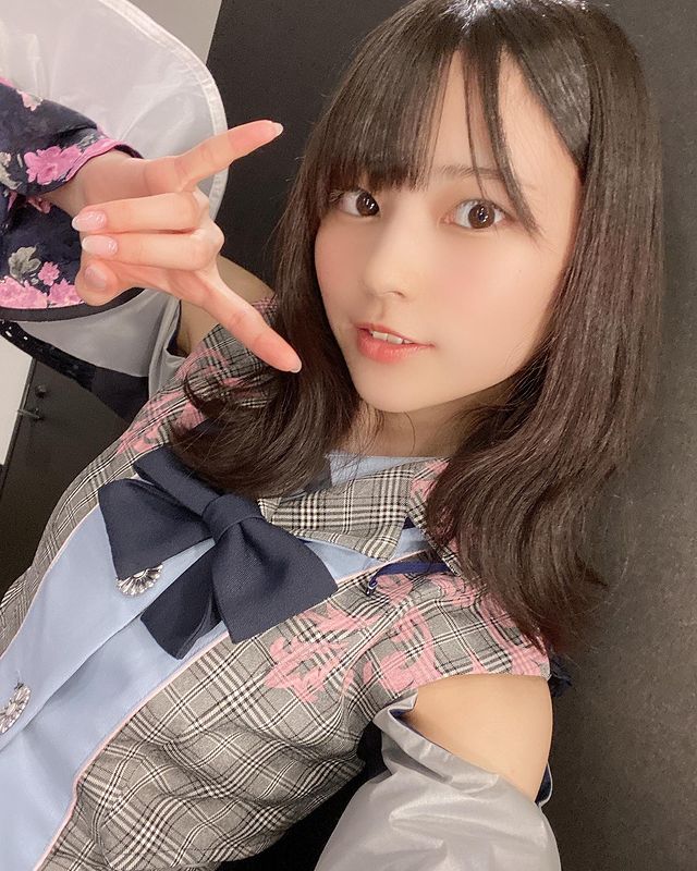 A young woman poses playfully for the camera, wearing a stylish outfit with floral accents. She is making a hand gesture while smiling, showcasing her youthful beauty and charm.